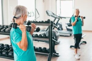 Smiling and active senior woman looking at herself in the gym mirror doing exercises with dumbbells. Sport, health, gym, wellness concept